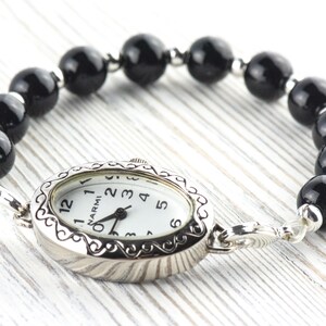 May include: A silver wristwatch with a white face and black hands, attached to a bracelet of large, glossy black beads. The watch face has black numerals and the brand name "NARMI".