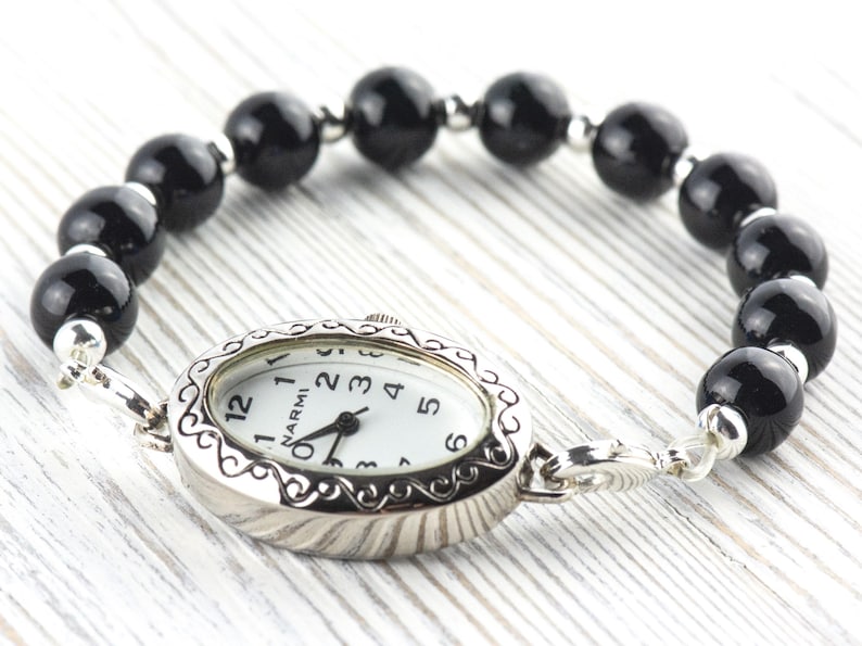 May include: A silver wristwatch with a white face and black hands, attached to a bracelet of large, glossy black beads. The watch face has black numerals and the brand name "NARMI".