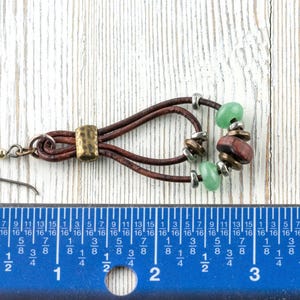 Pu&ograve; includere: Un orecchino fatto a mano con fili di cuoio marrone, impreziosito da perline verdi e rosso-marrone. Presenta un accento metallico color ottone e un gancio argentato. L'orecchino &egrave; lungo circa 5 cm.