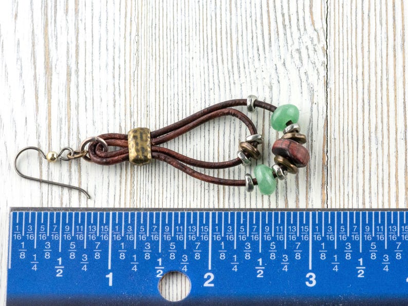 Pu&ograve; includere: Un orecchino fatto a mano con fili di cuoio marrone, impreziosito da perline verdi e rosso-marrone. Presenta un accento metallico color ottone e un gancio argentato. L'orecchino &egrave; lungo circa 5 cm.
