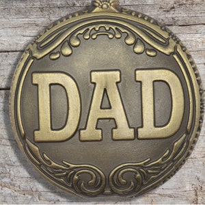 Peut inclure: Une d&eacute;coration ronde en m&eacute;tal dor&eacute; avec un motif orn&eacute; et le mot "DAD" en relief au centre.