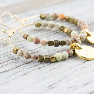 May include: A pair of gold hoop earrings with a beaded design. The beads are a mix of light brown, cream, and green colors. The earrings have a gold disc charm hanging from each hoop.