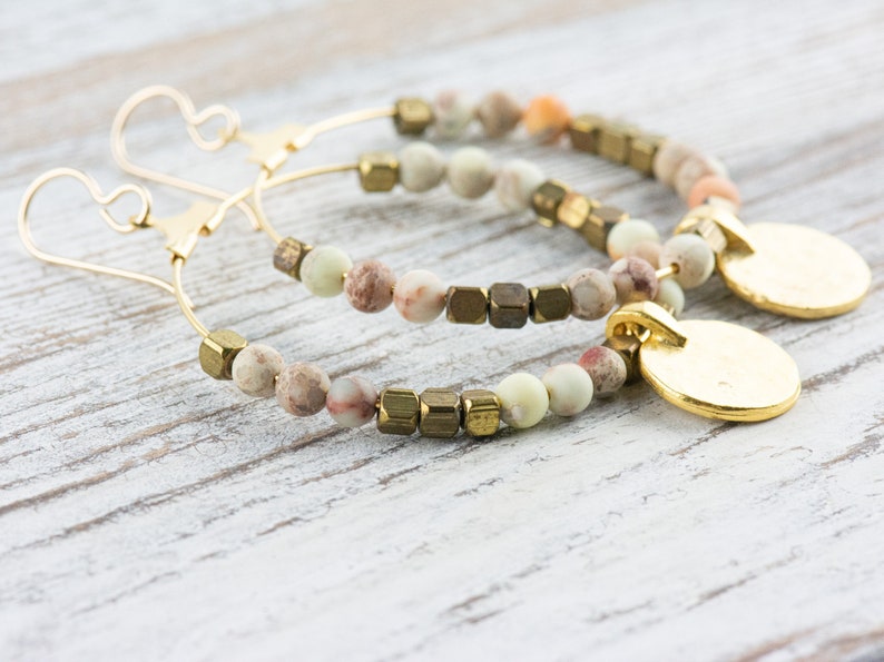 May include: A pair of gold hoop earrings with a beaded design. The beads are a mix of light brown, cream, and green colors. The earrings have a gold disc charm hanging from each hoop.