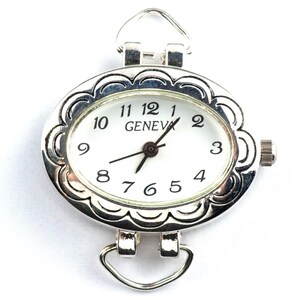 May include: Silver-coloured oval watch face with a white dial and black numerals. The watch face has a decorative scalloped edge and the word "GENEVA" is printed on the dial. It features a small crown and loops for attaching a strap.