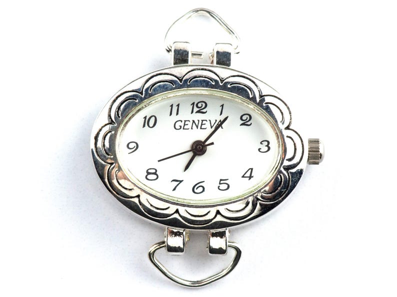 May include: Silver-coloured oval watch face with a white dial and black numerals. The watch face has a decorative scalloped edge and the word "GENEVA" is printed on the dial. It features a small crown and loops for attaching a strap.