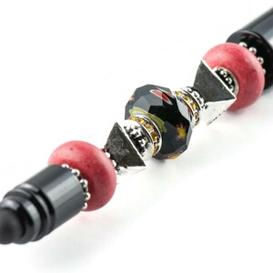 May include: A black stylus pen with a decorative design featuring red, black and silver beads. The beads are arranged in a pattern and have a sparkly finish.