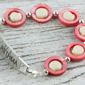 Pu&ograve; includere: Un braccialetto di allerta medica in argento con la parola "ASTHMA" incisa. Il braccialetto &egrave; decorato con perline di conchiglia rosa e bianca.