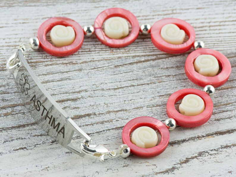 Pu&ograve; includere: Un braccialetto di allerta medica in argento con la parola "ASTHMA" incisa. Il braccialetto &egrave; decorato con perline di conchiglia rosa e bianca.
