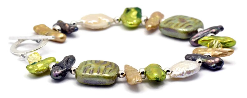 K&ouml;nnte beinhalten: Ein Armband aus gr&uuml;nen, braunen und wei&szlig;en Barockperlen mit silbernen Akzenten. Die Perlen sind unregelm&auml;&szlig;ig geformt und haben ein nat&uuml;rliches, organisches Aussehen.