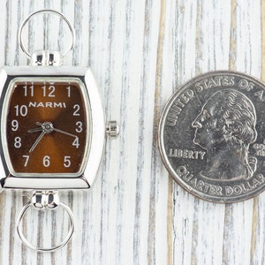 Puede incluir: Un reloj plateado con una esfera marr&oacute;n y n&uacute;meros negros. El reloj es aproximadamente del tama&ntilde;o de una moneda de 25 centavos de d&oacute;lar estadounidense.