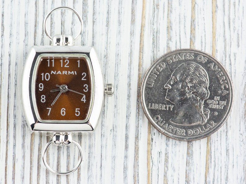 Puede incluir: Un reloj plateado con una esfera marr&oacute;n y n&uacute;meros negros. El reloj es aproximadamente del tama&ntilde;o de una moneda de 25 centavos de d&oacute;lar estadounidense.
