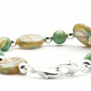 Op de afbeelding: Een zilveren armband met groene en bruine ovale kralen. De armband heeft een zilveren sluiting.