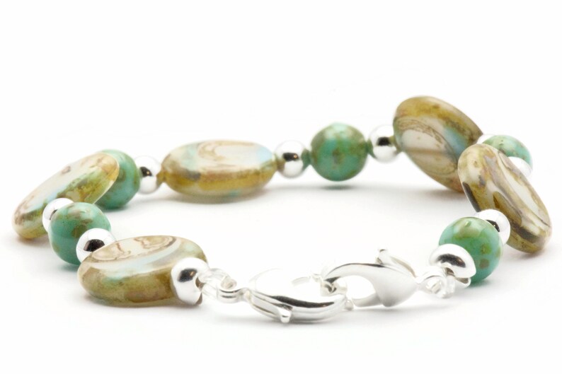 Op de afbeelding: Een zilveren armband met groene en bruine ovale kralen. De armband heeft een zilveren sluiting.