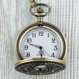K&ouml;nnte beinhalten: Antike Taschenuhr aus Messing mit wei&szlig;em Zifferblatt, schwarzen Ziffern und kunstvollem Filigran auf dem Deckel. Die Uhr zeigt die Zeit als zehn nach zehn. Das Wort "QUARTZ" ist auf dem Zifferblatt aufgedruckt.
