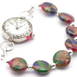 May include: A silver pocket watch with a white face and black hands attached to a bracelet made of colorful glass beads. The beads are oval shaped and have a gold, green, blue, and red speckled pattern.