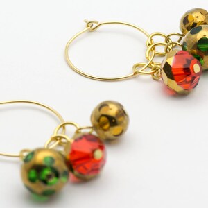 Puede incluir: Aretes de aro dorados con cuentas colgantes verdes, doradas y rojas. Las cuentas son redondas y tienen una superficie texturizada.