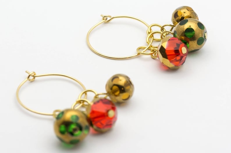 Puede incluir: Aretes de aro dorados con cuentas colgantes verdes, doradas y rojas. Las cuentas son redondas y tienen una superficie texturizada.