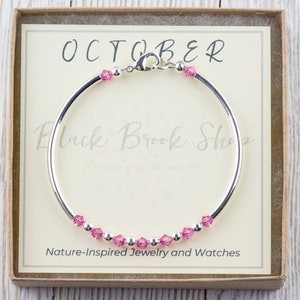 Puede incluir: Un brazalete de plata con cristales rosas y un cierre. El brazalete est&aacute; en una caja con el texto "OCTOBER" y "Black Brook Shop" en ella. La caja tambi&eacute;n dice "Joyer&iacute;a y relojes inspirados en la naturaleza".