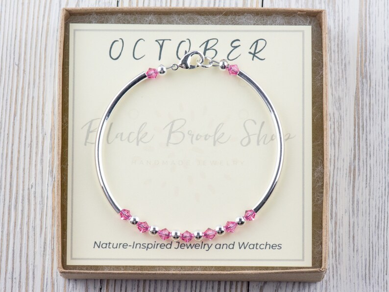 Puede incluir: Un brazalete de plata con cristales rosas y un cierre. El brazalete est&aacute; en una caja con el texto "OCTOBER" y "Black Brook Shop" en ella. La caja tambi&eacute;n dice "Joyer&iacute;a y relojes inspirados en la naturaleza".