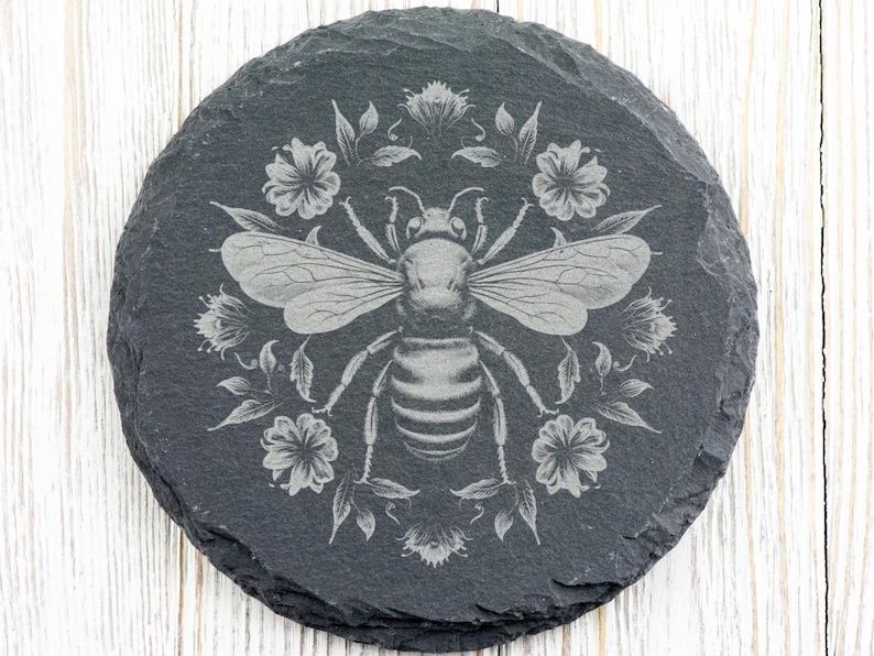May include: Round, dark gray slate coaster with an engraved design of a bee surrounded by flowers and leaves. The bee is centered, with detailed wings and body. The floral elements create a decorative border around the bee.
