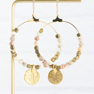 May include: Gold hoop earrings with a hammered gold disc charm. The hoops are adorned with white and pink marbled beads and gold cube beads.