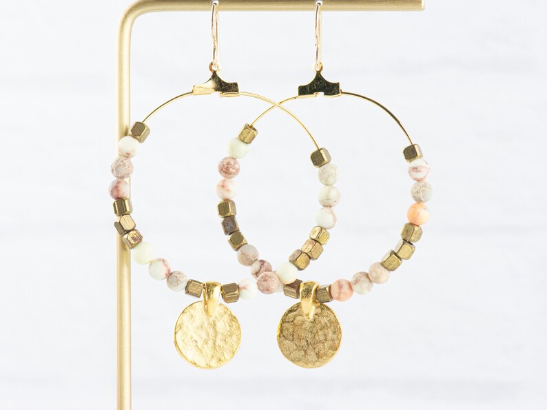 May include: Gold hoop earrings with a hammered gold disc charm. The hoops are adorned with white and pink marbled beads and gold cube beads.