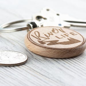 May include: A wooden keyring with a round, engraved design. The keyring features the text "River Life" with a stylised image of a river and trees.