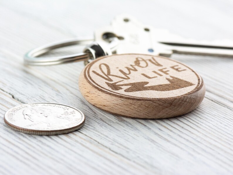 May include: A wooden keyring with a round, engraved design. The keyring features the text "River Life" with a stylised image of a river and trees.