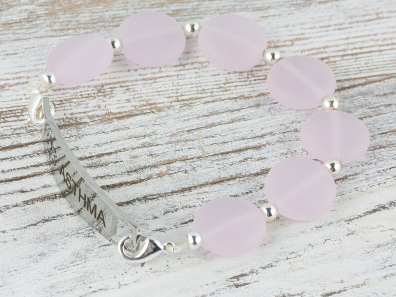 May include: A silver bracelet with a pink glass bead charm and a silver tag engraved with the word "ASTHMA".