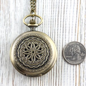 K&ouml;nnte beinhalten: Antike Taschenuhr aus Messing mit einem dekorativen Blumendesign auf dem Zifferblatt. Die Uhr ist rund und hat eine Kette. Eine M&uuml;nze liegt neben der Uhr zum Gr&ouml;&szlig;envergleich.