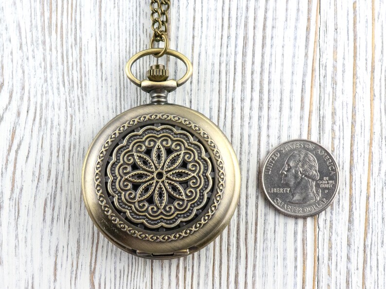 K&ouml;nnte beinhalten: Antike Taschenuhr aus Messing mit einem dekorativen Blumendesign auf dem Zifferblatt. Die Uhr ist rund und hat eine Kette. Eine M&uuml;nze liegt neben der Uhr zum Gr&ouml;&szlig;envergleich.