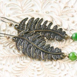 K&ouml;nnte beinhalten: Ein Paar Ohrringe mit einem Farnblatt-Design und einem gr&uuml;nen Stein-Akzent. Die Ohrringe sind aus Metall und haben eine rustikale, antike Oberfl&auml;che.