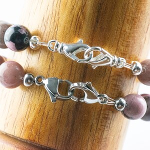 May include: Two beaded bracelets with silver clasps. The top bracelet has dark pink and black beads. The bottom bracelet has light pink beads.