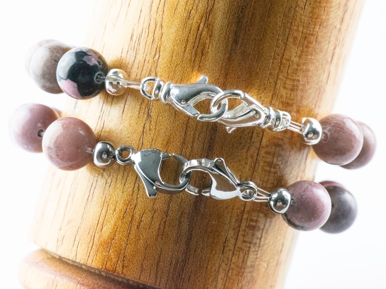 May include: Two beaded bracelets with silver clasps. The top bracelet has dark pink and black beads. The bottom bracelet has light pink beads.