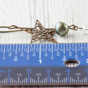 May include: A bronze-colored filigree star earring with a dark green pearl and a clear crystal bead. The earring is approximately 1.5 inches long, as measured by the blue ruler in the image. The earring is on a white wooden surface.
