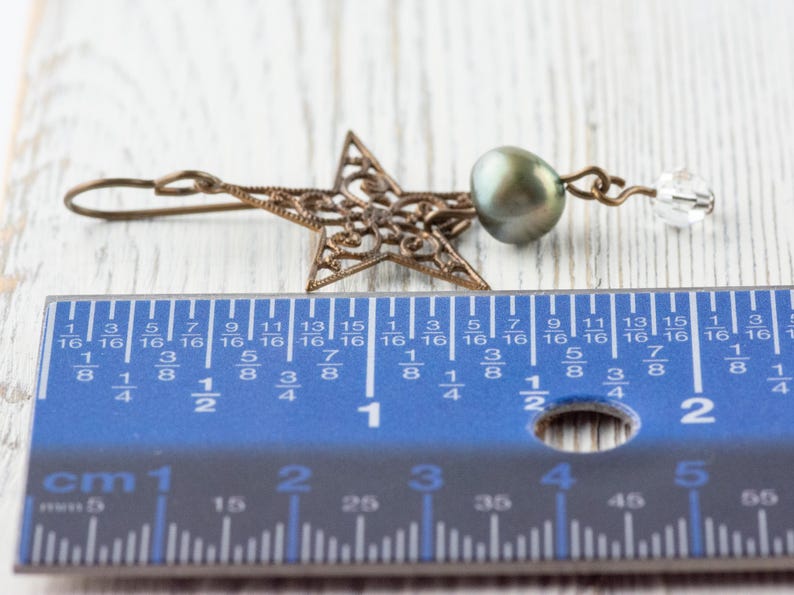 May include: A bronze-colored filigree star earring with a dark green pearl and a clear crystal bead. The earring is approximately 1.5 inches long, as measured by the blue ruler in the image. The earring is on a white wooden surface.