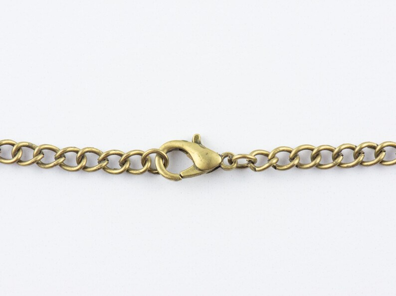 Op de afbeelding: Close-up van een messingkleurige ketting met een karabijnsluiting. De schakels van de ketting zijn ovaal van vorm en verbonden. De sluiting is open, klaar om bevestigd te worden. De achtergrond is effen wit.
