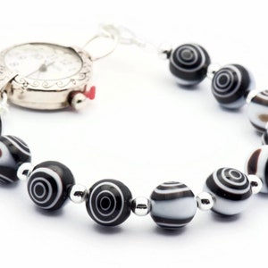 K&ouml;nnte beinhalten: Eine silberne Uhr mit wei&szlig;em Zifferblatt und schwarzen Zeigern, die an einem Armband aus schwarz-wei&szlig;en Perlen befestigt ist.