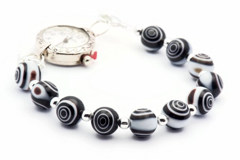 K&ouml;nnte beinhalten: Eine silberne Uhr mit wei&szlig;em Zifferblatt und schwarzen Zeigern, die an einem Armband aus schwarz-wei&szlig;en Perlen befestigt ist.