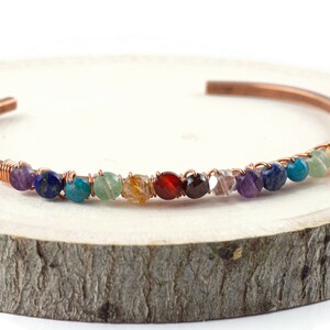 K&ouml;nnte beinhalten: Ein Kupferdraht-Wickelarmband mit einer Reihe von bunten Edelsteinen. Die Steine sind in der Reihenfolge des Regenbogens angeordnet: lila, blau, gr&uuml;n, gelb, orange, rot und klar.