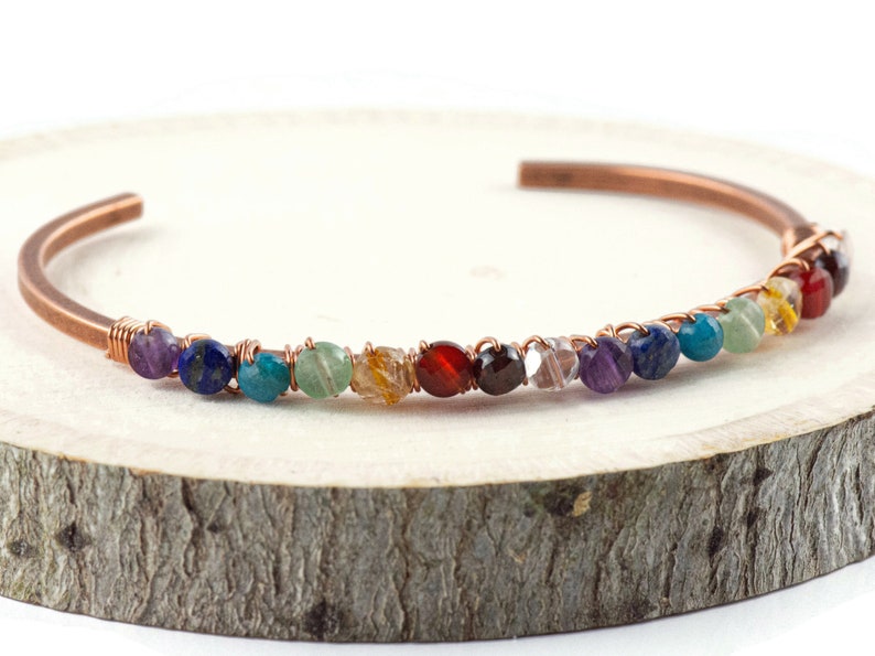 K&ouml;nnte beinhalten: Ein Kupferdraht-Wickelarmband mit einer Reihe von bunten Edelsteinen. Die Steine sind in der Reihenfolge des Regenbogens angeordnet: lila, blau, gr&uuml;n, gelb, orange, rot und klar.