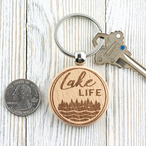 K&ouml;nnte beinhalten: Ein h&ouml;lzerner Lake Life-Schl&uuml;sselanh&auml;nger mit einem silbernen Schl&uuml;sselring und einem Schl&uuml;ssel. Der Schl&uuml;sselanh&auml;nger hat ein kreisf&ouml;rmiges Design mit den Worten &bdquo;Lake Life&ldquo; und einer Wald- und Wasserszene. Eine M&uuml;nze liegt neben dem Schl&uuml;sselanh&auml;nger.