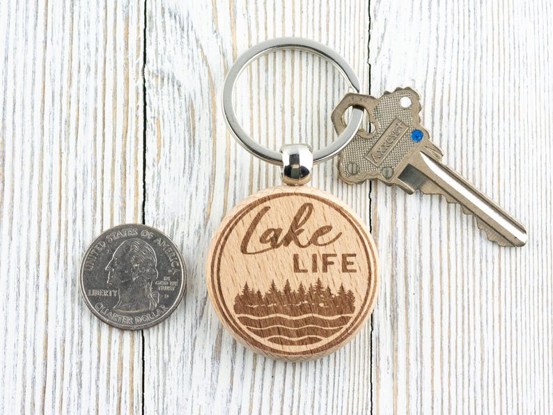 K&ouml;nnte beinhalten: Ein h&ouml;lzerner Lake Life-Schl&uuml;sselanh&auml;nger mit einem silbernen Schl&uuml;sselring und einem Schl&uuml;ssel. Der Schl&uuml;sselanh&auml;nger hat ein kreisf&ouml;rmiges Design mit den Worten &bdquo;Lake Life&ldquo; und einer Wald- und Wasserszene. Eine M&uuml;nze liegt neben dem Schl&uuml;sselanh&auml;nger.