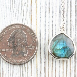 Pu&ograve; includere: Collana con pendente in argento con una pietra di labradorite sfaccettata a forma di goccia. La pietra mostra sfumature iridescenti blu e verdi. Una moneta da venticinque centesimi americana &egrave; accanto al pendente per il confronto delle dimensioni.