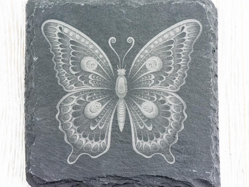 Puede incluir: Un posavasos de pizarra gris con un dise&ntilde;o de mariposa grabado en blanco. La mariposa tiene detalles intrincados y un patr&oacute;n delicado y sim&eacute;trico.