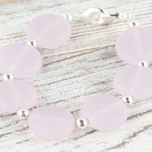 May include: A bracelet made of light pink oval-shaped beads with silver spacers. The bracelet is on a white wooden surface.