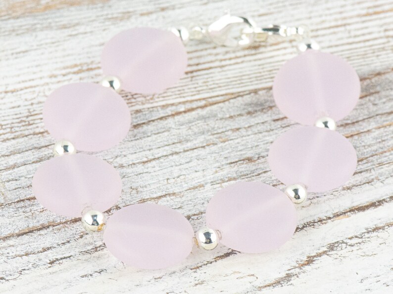 May include: A bracelet made of light pink oval-shaped beads with silver spacers. The bracelet is on a white wooden surface.