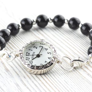 Pode incluir: Uma pulseira de rel&oacute;gio Geneva com mostrador branco e ponteiros pretos. O rel&oacute;gio est&aacute; ligado a uma pulseira de contas pretas e redondas. O rel&oacute;gio e a pulseira s&atilde;o de cor prateada.