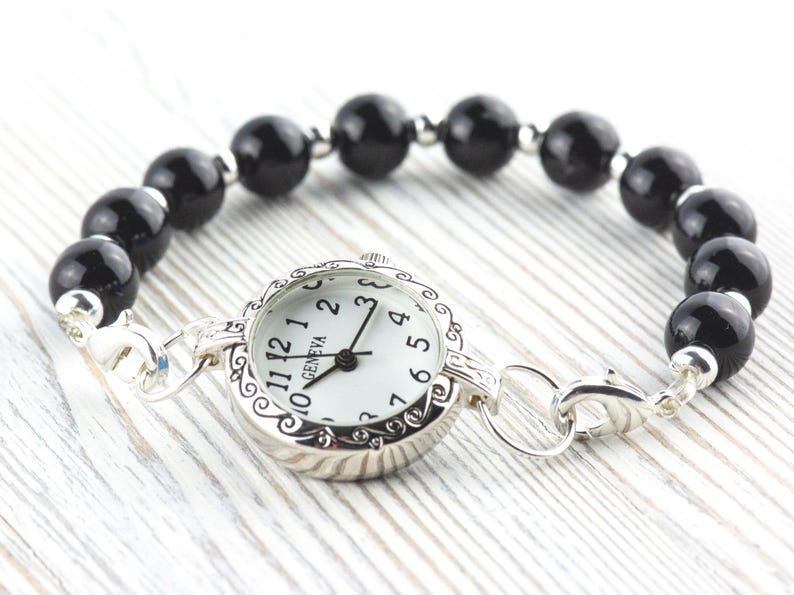 Pode incluir: Uma pulseira de rel&oacute;gio Geneva com mostrador branco e ponteiros pretos. O rel&oacute;gio est&aacute; ligado a uma pulseira de contas pretas e redondas. O rel&oacute;gio e a pulseira s&atilde;o de cor prateada.