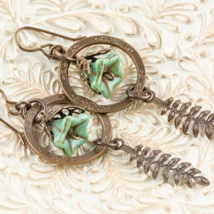 May include: A pair of handcrafted earrings with a bronze-coloured metal design. Each earring has a circular frame with a turquoise-coloured floral bead and a fern-shaped dangle. The earrings are displayed on a white, textured surface.